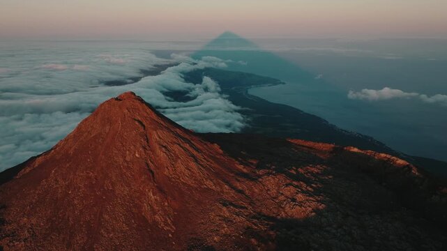 Breathtaking aerial perspective of the Mount Pico volcano summit at sunset, showcasing its perfect triangular shadow stretching over the Atlantic clouds and the island's coastline below.