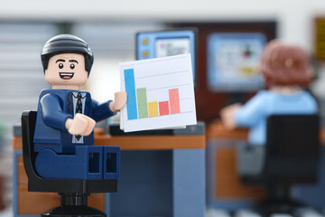 Obraz premium Tambov, Russian Federation - September 23, 2026 A happy Lego businessman minifigure sitting at his office desk, smiling and presenting a colorful bar chart document.