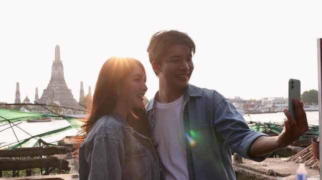 Happy young Asian couple taking selfie with smartphone, cheerful tourist man and woman enjoying city sightseeing trip near river temple landmark with Wat Arun, Bangkok, Thailand