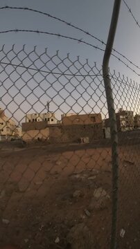 Old abandoned buildings seen through a chain-link and barbed wire fence at twilight.