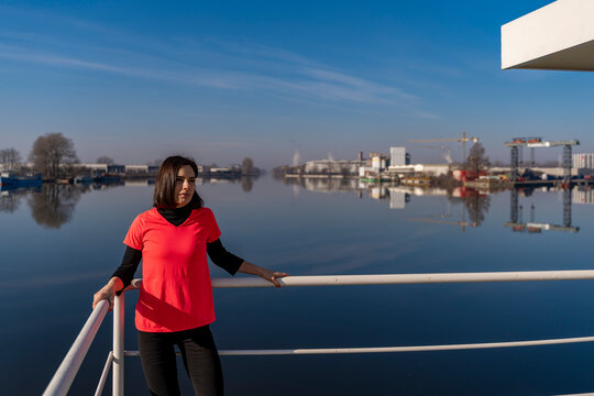 Woman standing by railing, looking at calm industrial river landscape with reflections under blue sky