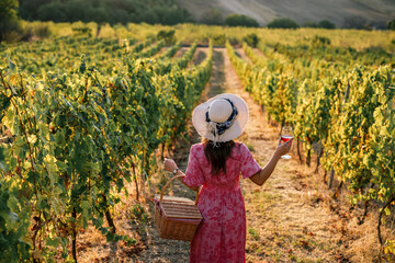 Obraz premium Woman with straw hat holding wine glass and basket walking in vineyard