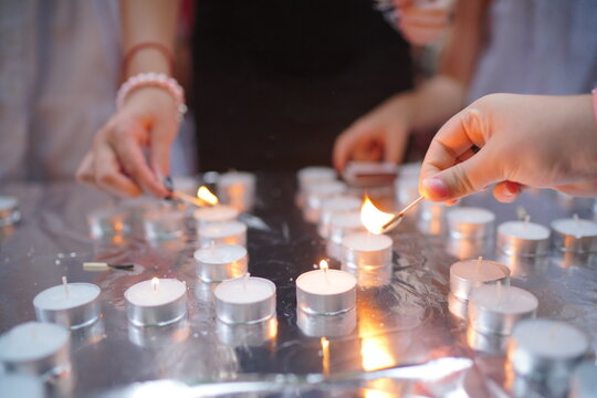 Hands light rows of tealight candles on a reflective surface during a quiet vigil, creating warm candlelight and an intimate atmosphere of remembrance, reflection and hope.
