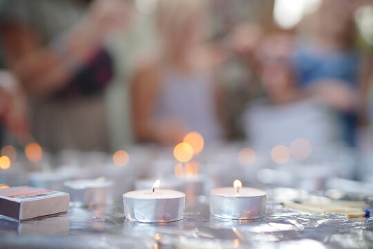 Softly glowing tea light candles sit on a reflective surface while a blurred group of people gathers in the background, evoking warm celebration, intimacy, and quiet festive atmosphere.