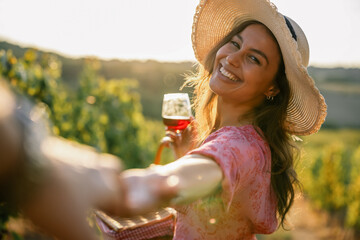 Obraz premium Woman holding hand leading partner through vineyard holding wine glass at sunset