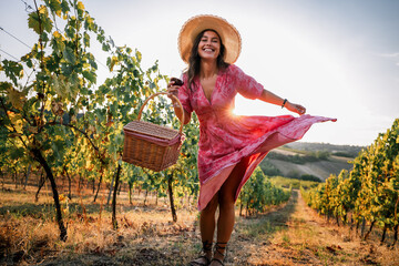 Obraz premium Playful woman twirling pink dress in vineyard holding picnic basket and wine