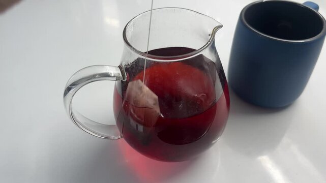 Close up of a hand pulling a tea bag from a clear glass pitcher filled with hot red herbal tea next to a blue mug