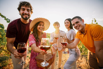 Obraz premium Cheerfully smiling friends holding out red wine glasses at vineyard picnic