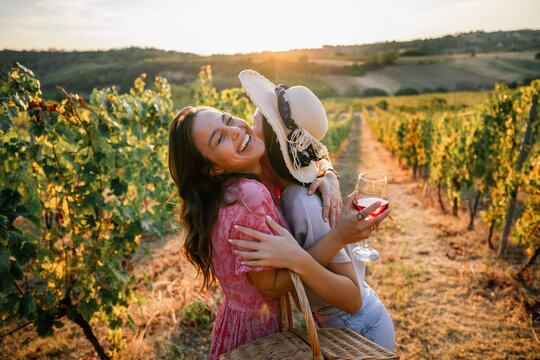 Two joyful female friends hugging tightly with wine in vineyard sunset