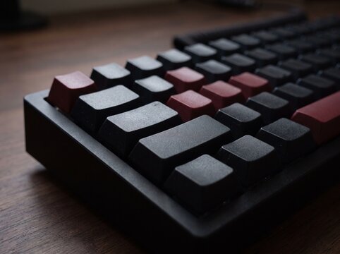 A close-up, low-angle shot of a mechanical keyboard with dark grey and red keycaps on a wooden desk.