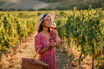 Obraz premium Woman in straw hat smelling glass of red wine in vineyard at sunset