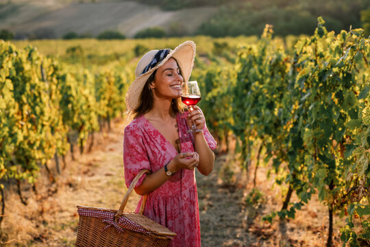 Woman in straw hat smelling glass of red wine in vineyard at sunset