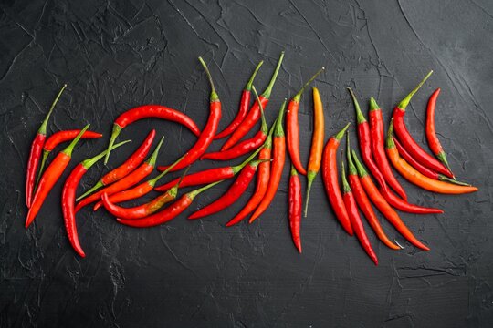 Bright red and orange cayenne chili peppers arranged in a scattered cluster on dark textured stone surface with green stems visible under even top-down lighting