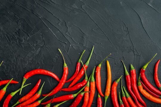 Ripe red and orange cayenne chili peppers arranged in clusters on a dark textured stone surface with green stems visible under even overhead lighting
