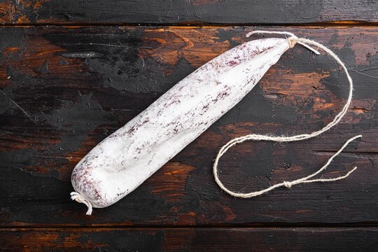 White powdery coated dry cured spanish longaniza sausage resting on dark rustic wooden planks with natural wood grain texture, tied at ends with coarse twine, under even top-down lighting