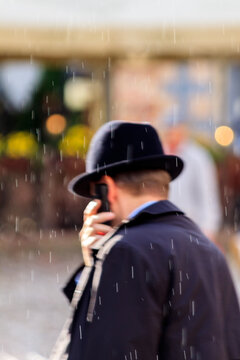 A man in a black bowler hat and dark coat stands with his back to the camera in falling rain in Tallinn, Estonia, blurred lights and figures visible in the softly focused background