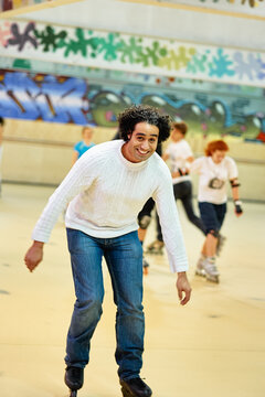 Smiling man skating at indoor roller rink with friends in the background, conveying fun, leisure and social activity. Casual clothing, energetic movement and colorful graffiti set playful urban mood.