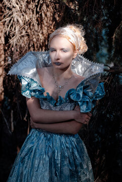 A dramatic fantasy portrait of an ice queen in a shimmering blue gown and silver collar, posing with folded arms in a shadowed forest. Cold makeup, elegant jewelry and mysterious, regal mood.