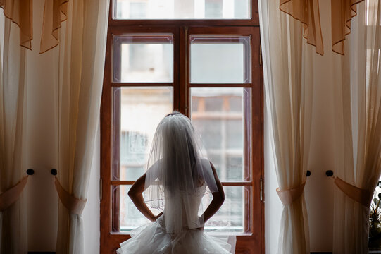 A bride in a tulle gown and veil stands with hands on hips, gazing out a vintage window in a softly lit bridal suite, capturing a quiet, contemplative moment before the wedding.