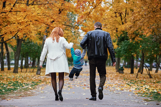 Parents hold their young child by the hands and swing him playfully along a tree-lined park path covered in golden autumn leaves, capturing warm family bonding and joyful outdoor moments.