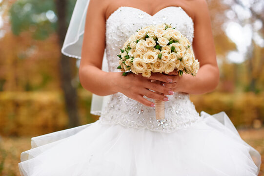 A bride in a beaded white wedding dress holds a cream rose bridal bouquet while standing outdoors among autumn foliage, evoking romance, celebration, elegance, and intimate wedding moments.