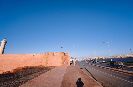 Lighthouse, pink wall, clear blue sky, and peaceful road evoke serene coastal scenery. Historic Coastal Road With Lighthouse and Pink Wall Under Clear Blue Sky