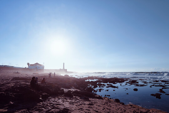 A serene coastal setting featuring a lighthouse, a nearby house, and people relaxing on the shore. The bright sun enhances the tranquil atmosphere and emphasizes the beauty of the ocean.