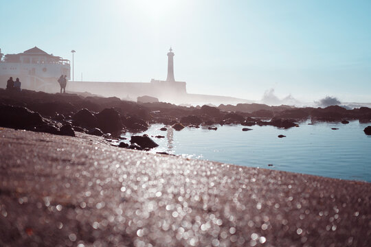 A peaceful coastal scene featuring a lighthouse, waves, and shimmering sunlight reflecting on wet rocks.