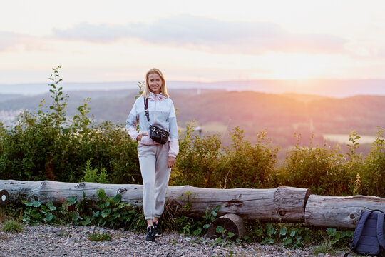 A smiling young woman in casual athleisure stands by a fallen log overlooking a sunlit valley at dusk, enjoying peaceful nature, fresh air and a scenic hiking viewpoint.