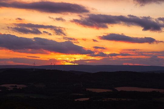 Vibrant orange and purple sunset lighting a layered landscape of rolling hills and silhouetted wind turbines, evoking renewable energy, calm evening atmosphere and dramatic natural beauty.