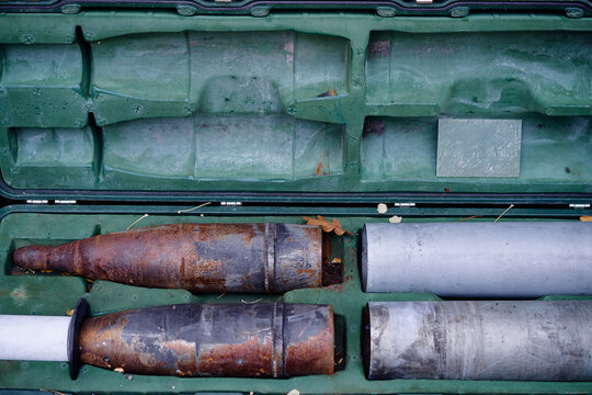 Weathered artillery shells and cylindrical metal casings resting in a green foam-lined carrying case, showing rust, wear and industrial texture on vintage military munitions and equipment.
