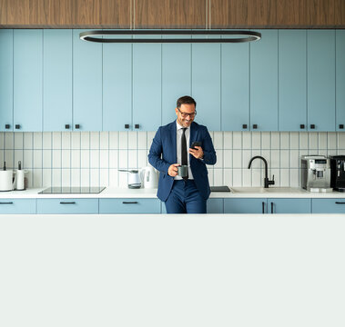 Businessman in an office kitchen space checking messages on his mobile phone and holding a coffee mug, enjoying a quick break from work