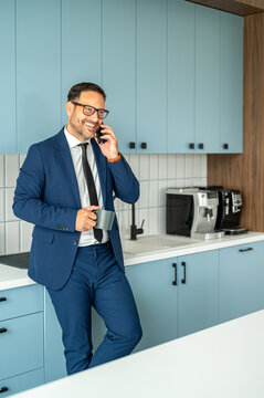 Professional man discussing business on phone in office kitchen, smiling and enjoying coffee during break, communicating with clients, colleagues, or partners