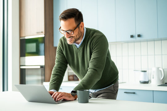 Smiling man in glasses working on a laptop at a modern kitchen table, focused on digital tasks like bookkeeping or legal consulting, showing productive remote work life