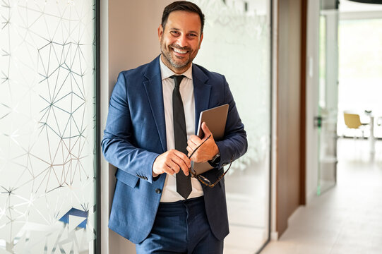 Professional businessman holding glasses and digital tablet, smiling confidently at the camera in a modern office, representing corporate success and expertise