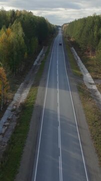 Drone footage of a long straight asphalt highway cutting through colorful autumn forest, with a single car driving in the distance, Ural region, Russia