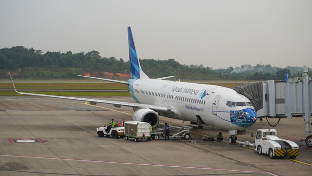 Garuda Indonesia Boeing 737-800 with special face mask livery parked at Hang Nadim International Airport tarmac connected to jet bridge during ground handling.