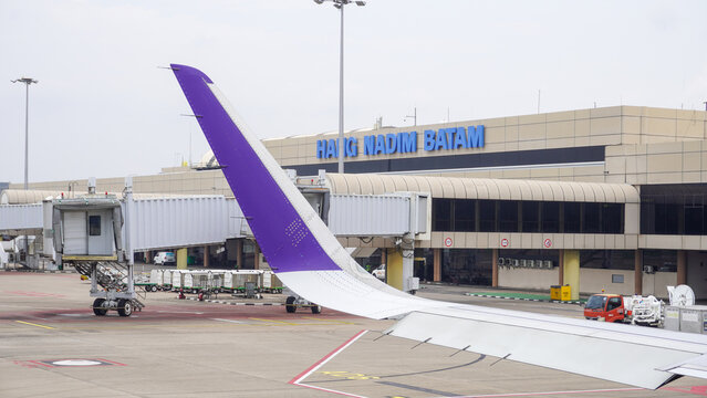 Airplane wing parked on the tarmac with Hang Nadim International Airport terminal building in Batam, Indonesia. Boarding bridge and ground service vehicles background for travel and aviation.