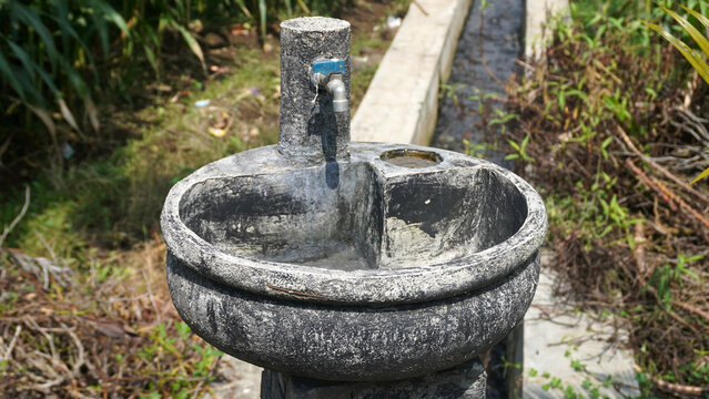 Weathered stone drinking fountain with a small faucet, water flowing into a round basin surrounded by greenery.