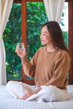 Vertical portrait image of a woman holding and drinking water on the bed at home