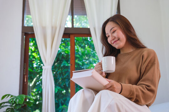 Portrait image of a woman drinking coffee and reading a book on the bed at home