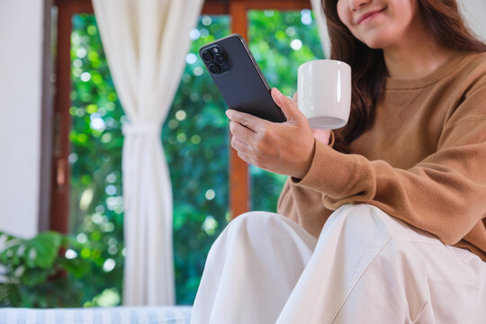 Closeup image of a woman holding and using smart phone while drinking coffee on the bed at home