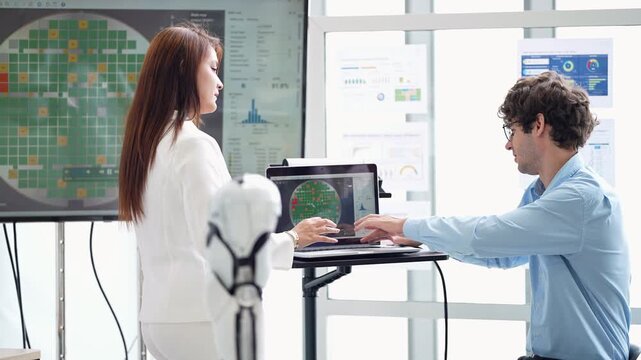 Engineer in blue shirt types on a laptop cart while a woman in white suit touches her hair and follows the circular wafer scan results with him, robotics R and D