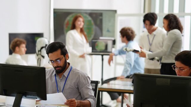 Bearded engineer reviews papers at his desk while teammates test a bionic hand and discuss screens in a busy tech office, during an iterative design check, a tech startup innovation concept