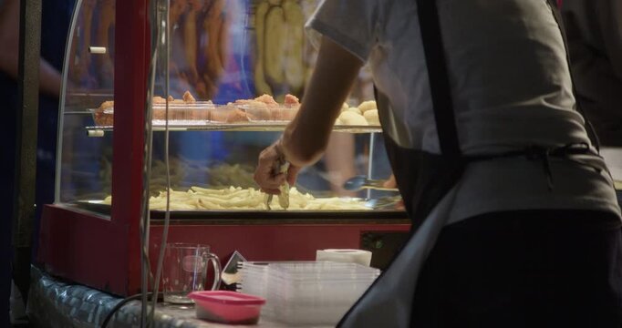 Vendor using tongs to pick cooked food from a glass display case at a night market stall. Close-up of ready-to-eat streetfood selection and serving in an outdoor setting.