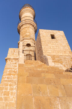 Historic Venetian lighthouse and stone harbor wall illuminated by warm sunrise light in Chania, Crete, Greece.