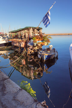 Colorful floating souvenir shop with Greek flag and small boats reflected in calm water of Chania harbor, Crete, Greece.