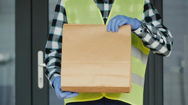 Delivery courier wearing safety vest and gloves holding brown paper bag for takeaway food shipment standing outdoors.
