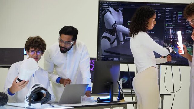 Bearded engineer in goggles lifts a white robotic forearm shell to show a colleague at the laptop as a curly-haired woman in an exoskeleton arm reads the simulation screen