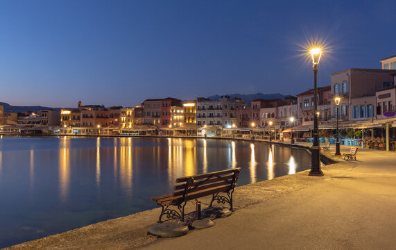 Waterfront buildings and calm harbor water with illuminated street lamps before sunrise in Chania, Crete, Greece.